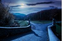 park among mountain in autumn at night in full moon light. trees in colorful foliage, vivid grassy green lawns. walking path to the bridge through creek