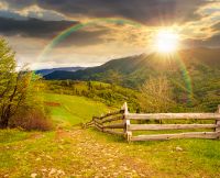 rolling hills of rural landscape in green grass at sunset. wooden fence in mountains in evening light. alpine countryside in spring. cloudy sky. hope, expectation, new beginning and bright future