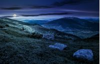 huge stones among the grass on top of the hillside meadow near the edge of a mountain. vivid summer landscape at night in full moon light