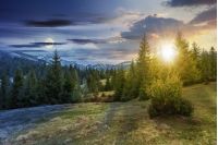 day and night time change concept of beautiful springtime landscape with sun and moon. spruce forest on grassy hillside meadow. spots of snow on distant ridge. 