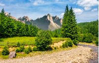 composite summer landscape with trees on a cliff nearthe shore of a clear river at the foot of epic High Tatra mountain ridge with rocky peaks under blue sky with clouds